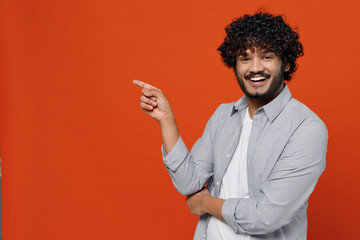 Smiling fascinating happy young bearded Indian man 20s years old wears blue shirt pointing index finger aside on workspace area copy space mock up isolated on plain orange background studio portrait.