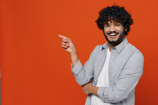 Smiling Fascinating Happy Young Bearded Indian Man 20s Years Old Wears Blue Shirt Pointing Index Finger Aside On Workspace Area Copy Space Mock Up Isolated On Plain Orange Background Studio Portrait.