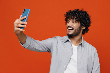 Obraz na płótnie Canvas Fun fascinating friendly young bearded Indian man 20s years old wear blue shirt doing selfie shot on mobile cell phone post photo on social network isolated on plain orange background studio portrait