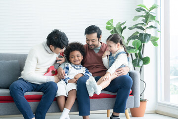Asian young LGBTQ gay couple playing ukulele and singing with little Caucasian and African adopted kid together at home. Happy son and daughter smiling and sitting on sofa. LGBT diverse family concept