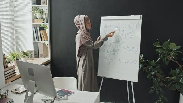 Muslim teacher in hijab explaining math formulas on flipchart while giving online lesson via video call on computer
