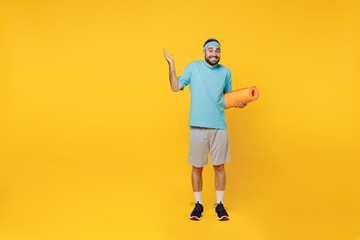 Fototapeta na wymiar Full body young fitness trainer instructor sporty man sportsman in headband blue t-shirt spend weekend in home gym hold yoga mat spread hands isolated on plain yellow background Workout sport concept