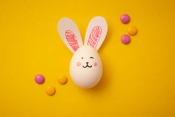 Food photo for Happy Easter. Chicken eggs with cute bunny faces and bunny ears and yellow and pink candies on a yellow background. Preparation for the holiday. Greeting card for Easter holiday.