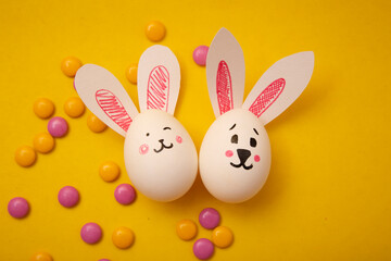 Food photo for Happy Easter. Chicken eggs with cute bunny faces and bunny ears and yellow and pink candies on a yellow background. Preparation for the holiday. Greeting card for Easter holiday.