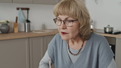 Tilt up shot of senior woman writing down notes and watching online lesson on laptop while learning remotely at home