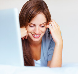 Happy woman in front of laptop on bed and speaking on cellphone. Cute Caucasian woman speaking on cellphone , lying in front of laptop in bed.