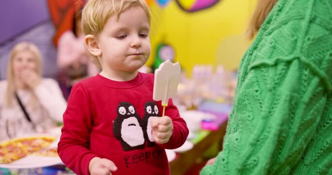 A little birthday boy bites a candy on a stick from a birthday cake. Boy surrounded by family and friends. Birthday party
