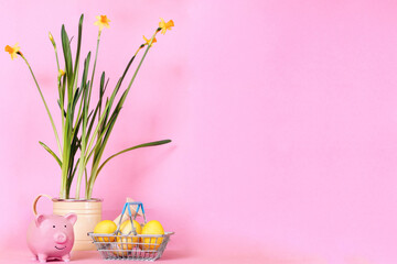 eggs in an iron grocery basket daffodils in a pot watering can piggy bank on a pink background. Savings at Easter, discounts and sales. copyspase, a place for text