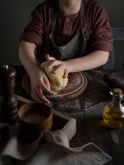Kid making pelmeni (meet dumplings) of dough on wooden table with ingredients flour, oil, salt, dark background. Copy space. Home bakery concept, kitchen cooking story.