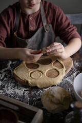 Kid making pelmeni (meet dumplings) of dough on wooden table with ingredients flour, oil, salt, dark background. Copy space. Home bakery concept, kitchen cooking story.