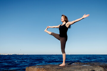 Obraz premium Woman doing yoga background blue sky and sea. Tree pose or vrksasana with hands together up above head. Concept relaxation, harmony with nature