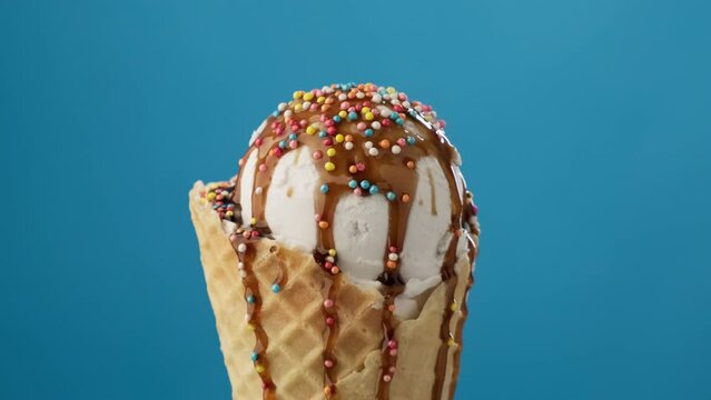 Vanilla white ice cream rotating at blue background. Ice cream covered with caramel or chocolate topping and colorful sugar sprinkles. Close-up of delicious dessert in waffle cone. 4K, UHD