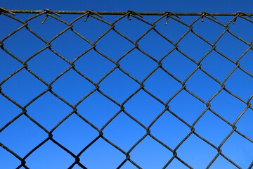 Fototapeta premium Gray or dark metal fence chain link. Close up and isolated. Part of a greater fence. Clear blue sky in the background.