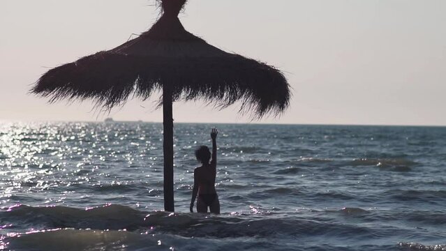 Silhouette of attractive woman standing under the straw umbrella in the sea and waving to the boat on the horizon, lost lonely woman, slow motion