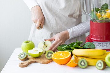 Young woman preparing homemade vitamin smoothie with apples, spinach, banana, kiwi, orange. Female hands. Process of preparation healthy food. Concept of clean eating, dieting, low calories food.