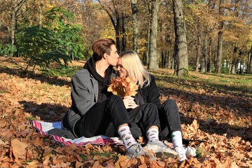 A young couple in a coat sit in a park or forest. A couple in love - a blonde girl and a guy with blond hair. The young man hugs and kisses the girl. Joint walk. Romantic relationship. Valentine's Day