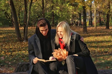 A young couple in a coat sit on a log and read a book together. A couple in love - a blonde girl and a guy with blond hair. Joint walk in the park or forest. Romantic relationship. Valentine's Day.