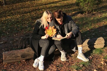 A young couple in a coat sit on a log and read a book together. A couple in love - a blonde girl and a guy with blond hair. Joint walk in the park or forest. Romantic relationship. Valentine's Day.