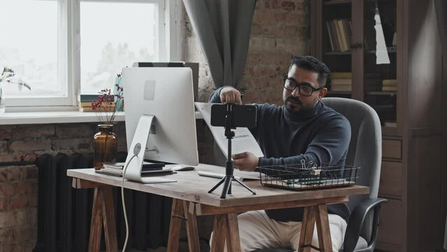 Middle eastern male teacher sitting at desk in home office and explaining task in workbook on smartphone camera on tripod while giving online lesson