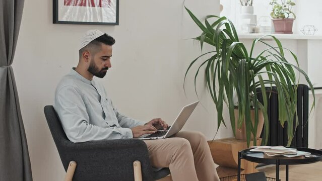 Muslim Man In Kufi Sitting In Armchair At Home And Typing On Laptop While Working Remotely From Home