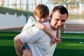 Side view of smiling father carrying cheerful boy on back while spending time together at football field