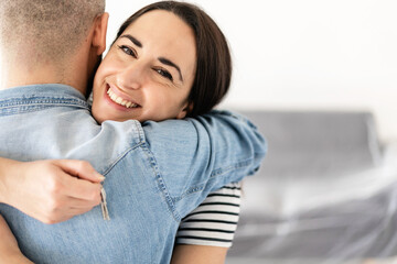 Happy young couple in their new apartment - 30s woman holding new apartment keys while hugging her boyfriend in the living room - Relocation and moving concept