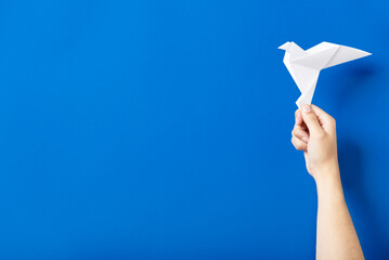 Hand holding a white paper dove, symbol of peace, against a blue background.