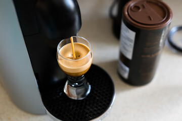 Fresh homemade cup of espresso filling in a generic coffee machine. Modern espresso machine on kitchen table and glass filling with coffee crema