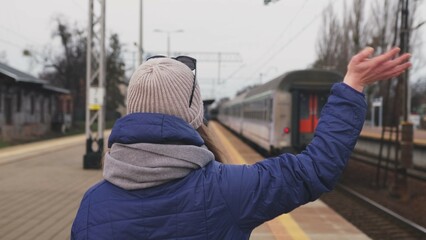 Young Caucasian Female Standing on Platform Waving Hand Bidding Farewell To Loved One Relative Leaving on Outbound Passenger Train Departing Railway Station	