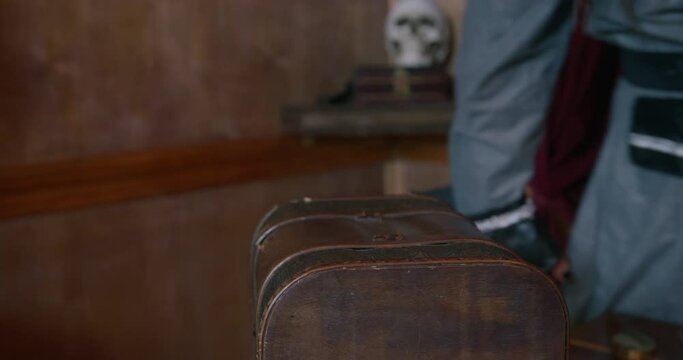 Pirate captain in long coat knocks on brown treasure chest standing on table IN cabin of pirates ship during voyage close view