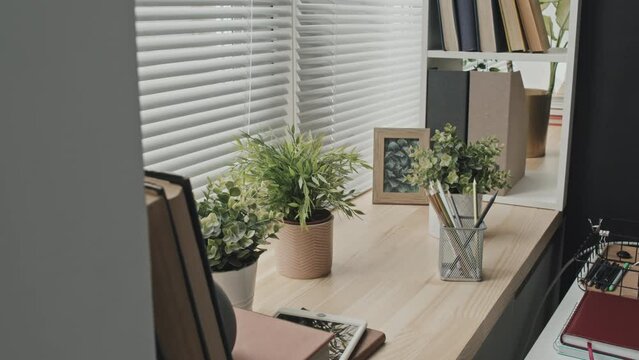 Arc shot of houseplants, digital tablet, books, framed picture, notepad and pencils in mesh holder on wooden window sill in home office