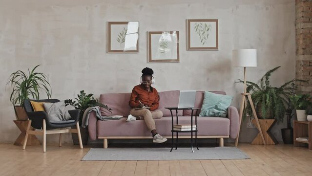 Wide shot of young African American woman in wireless headphones sitting on sofa in living room, watching online class on laptop and taking notes while studying from home
