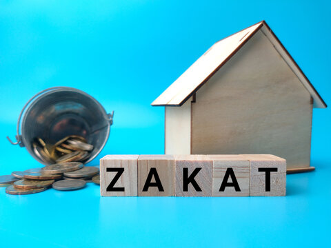 Wooden House And Coins With Text ZAKAT On A Blue Background.