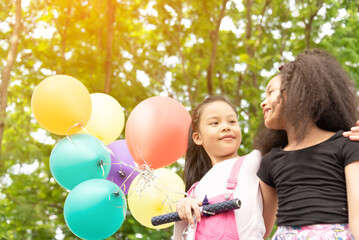 Fototapeta na wymiar Happy little asian & african american girl playing colorful balloons together. Trees and green gardens background. Smiling lovely girl with balloons on the street in the summer. 