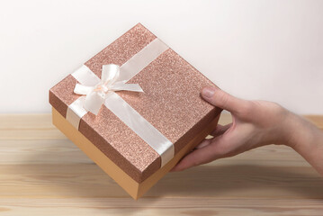 A hand gives a gift over a wooden table. The hand holds a pink gift box tied with a white satin bow.
