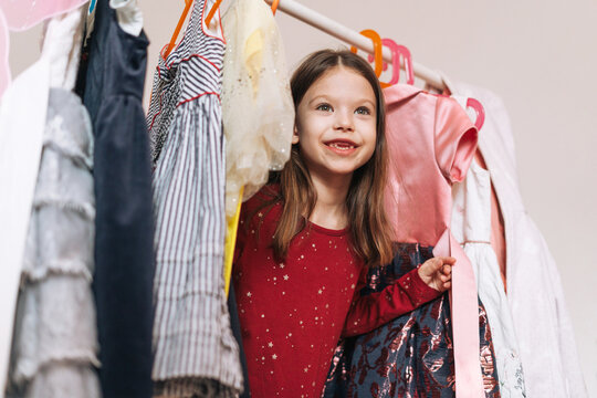 Smiling little girl with long dark hair in red dress among her beautiful dresses in wardrobe in children's room at home