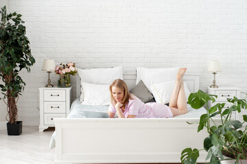 Obraz na płótnie Canvas A pleasant blonde woman in pajamas lies on a bed in a bedroom with white walls and houseplants and holds a laptop in her hands. Work from home. Online shopping.