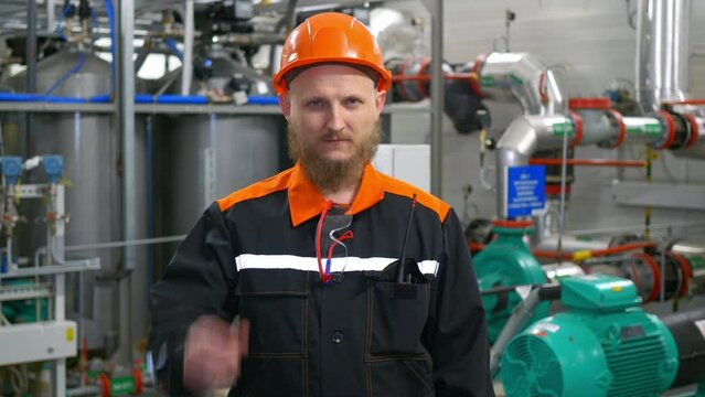 A bearded man is a technological specialist in an orange helmet, the operator of the technological process of the production line of a gas compression plant at his workplace.