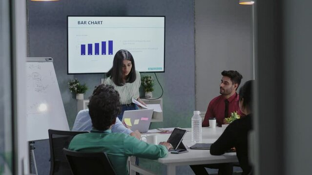 Young Indian female business trainer company manager coach wear suit presenting explain design project result give flip chart presentation training business team group at office workshop meeting.