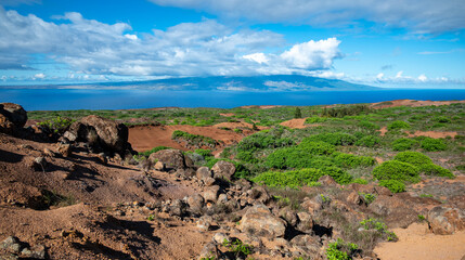 Looking across a rugged landscape  on Lanai of red dirt, growing shrubs and boulders to the ocean and the island of Maui across the channel.
