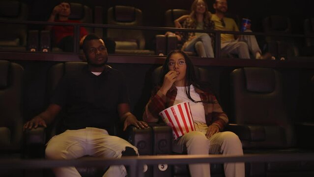 African American young couple sitting in movie theatre watching film and eating popcorn