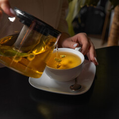 Close up of tea from the kettle slowly pour into porcelain cup. Young lady preparing green tea in the kitchen in the morning