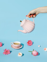 Creative layout with hand holding teapot and pouring tea into pink cup with with fresh flowers...