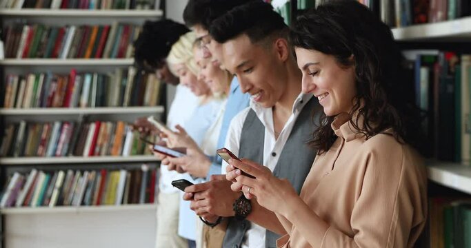 Group of happy diverse young people of different ethnicities cultures stand in line close to library bookshelves use phones. Gen z international university students engaged in work with gadgets online