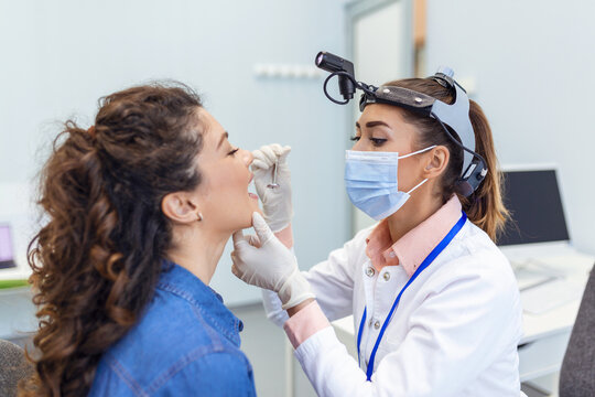 A Young Woman Sits On An Exam Table Across From Her Doctor. The Doctor Reaches Forward With A Tongue Depressor As The Woman Looks Up And Sticks Out Her Tongue.