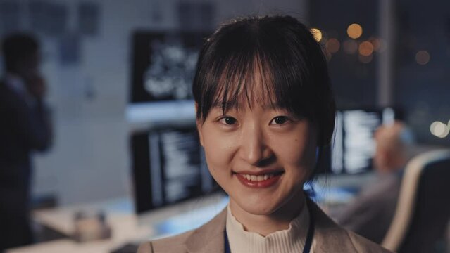Close-up of young Asian woman dressed formally, standing in corporate office at night, smiling and looking on camera, blurred colleagues working on background