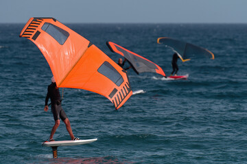 A group is wing foiling using handheld inflatable wings and hydrofoil surfboards in a blue ocean,...