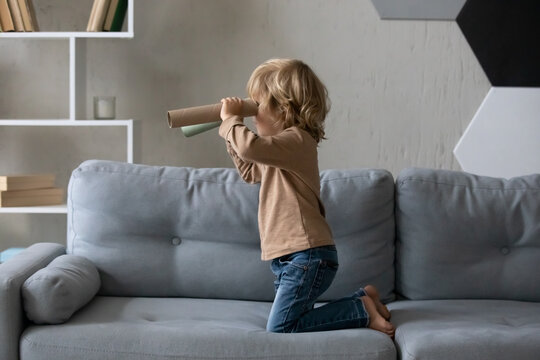 Happy adorable small child boy playing pirates game, looking in distance using carton spyglasses, having fun on huge comfortable couch alone at home, hobby adventure playtime activity concept.