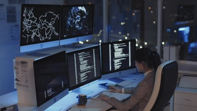 Side view of young Asian female programmer sitting at desk in corporate office at night, coding, using multiple monitors