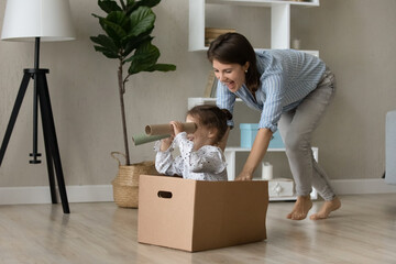 Joyful laughing young mother pushing cardboard box with seated little adorable child daughter...